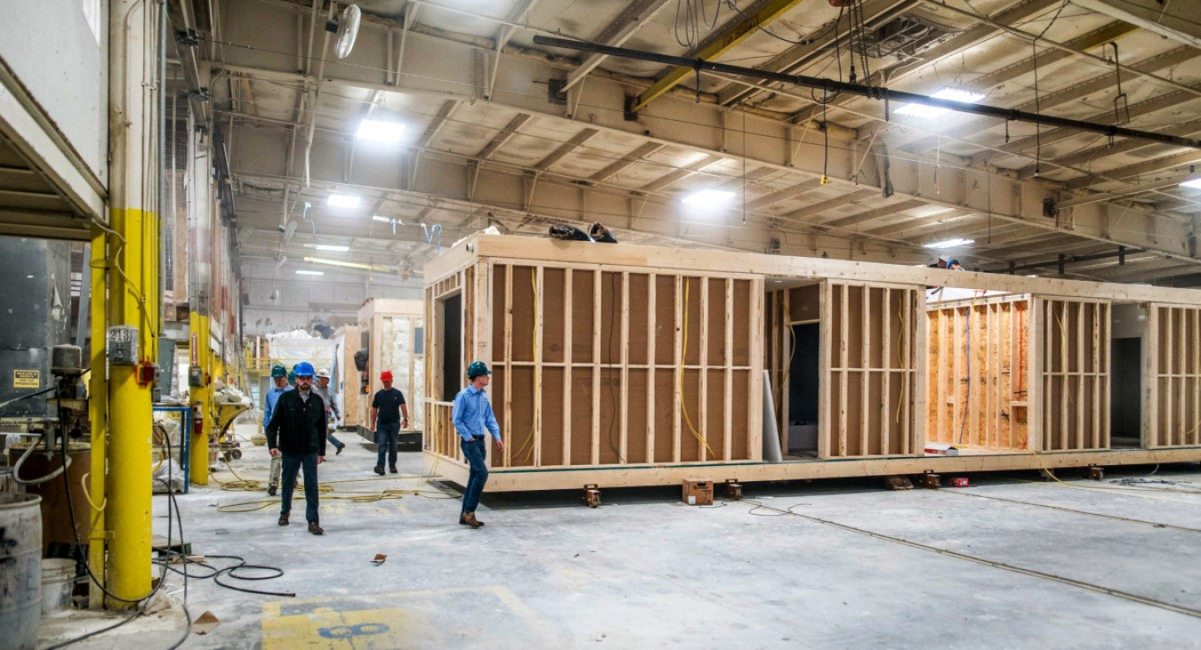 The inside of a large warehouse with a construction project going on and workers walking around it.