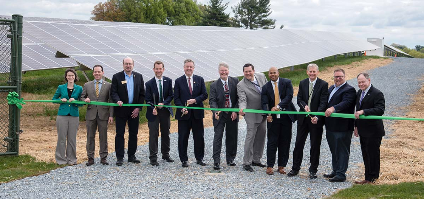 Image of 10 men and one woman holding a green ribbon with three men in the middle holding scissors to cut the ribbon.