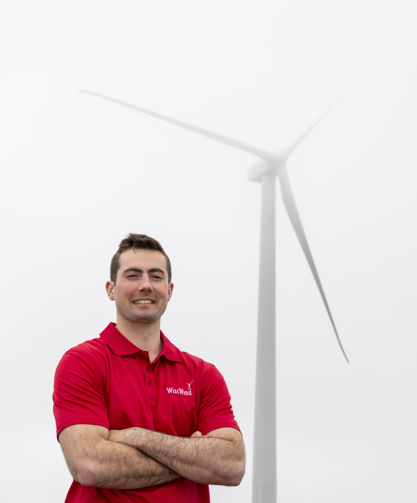 A student in a red shirt stands in front of a wind turbine on a cloudy day.