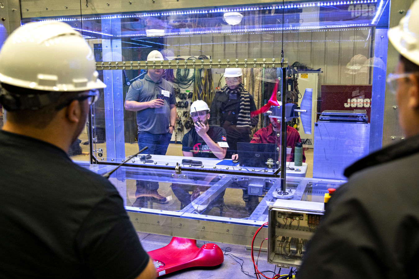 Three students wearing safety gear are visible through a glass enclosure with a small-scale wind turbine, while two people in the foreground take measurements.