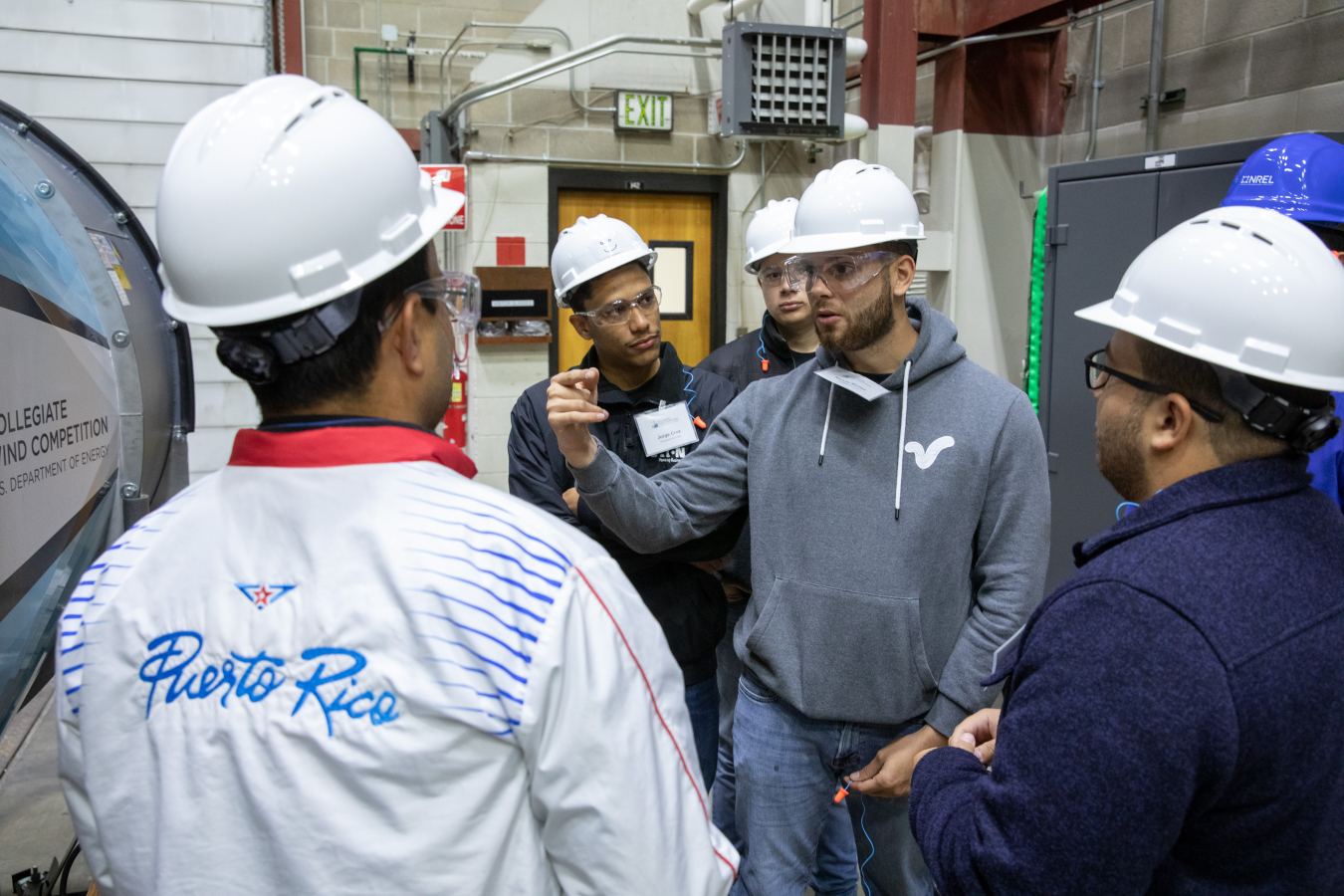 Five students in safety gear stand in a huddle next to a wind tunnel.