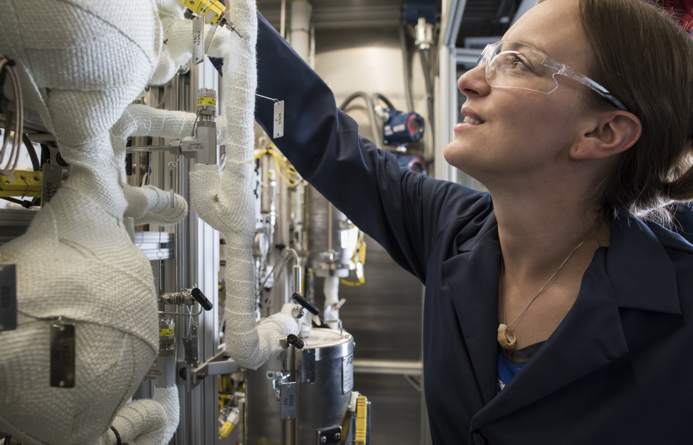 Female researcher in blue lab coat and safety glasses adjusting scientific equipment with wrapped components mounted vertically in a case.