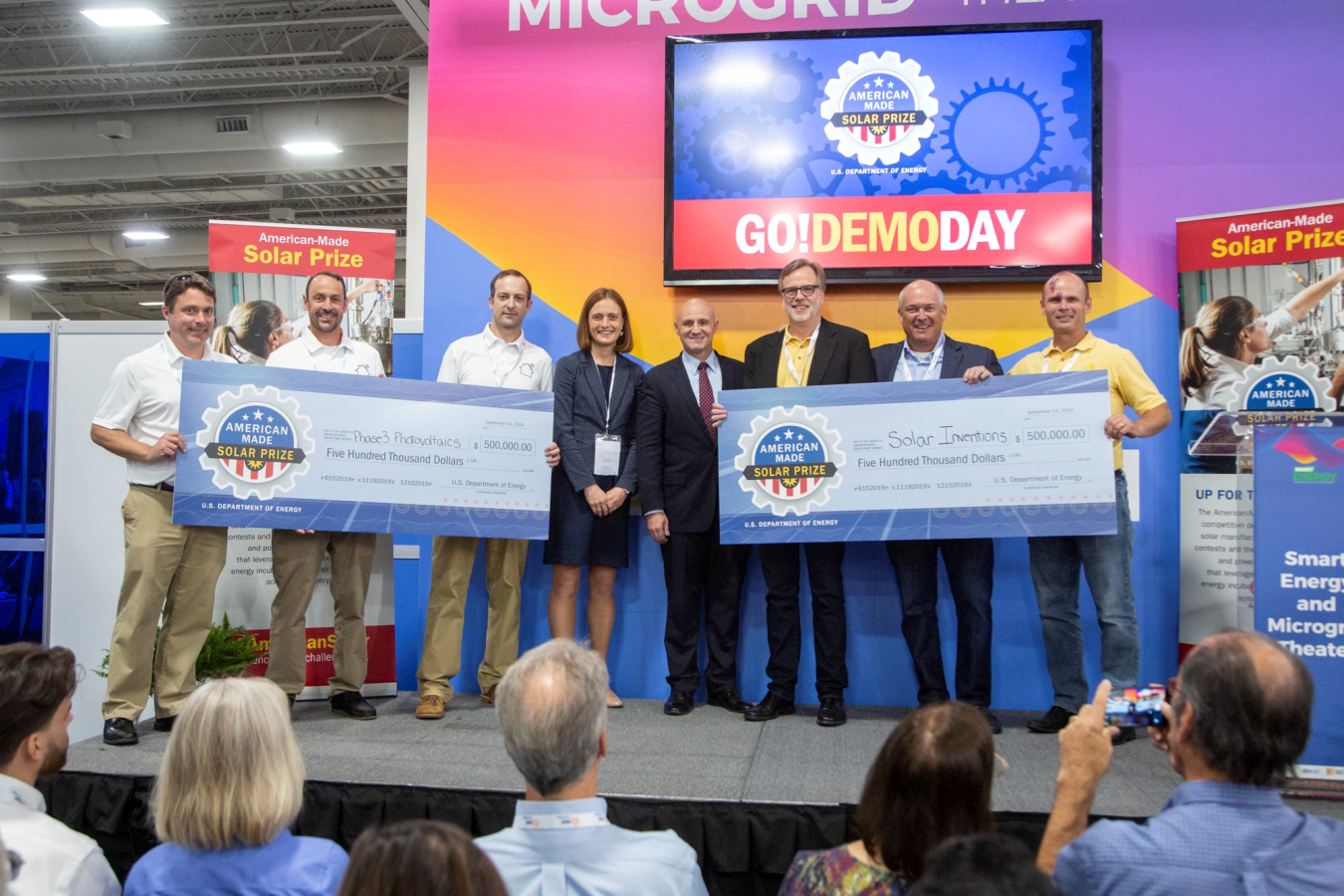 Three winners from Phase 3 Photovoltaics and three from Solar Inventions hold up mock checks while Becca Jones Albertus of the Solar office and Assistant Secretary Daniel Simmons stand in the middle. 