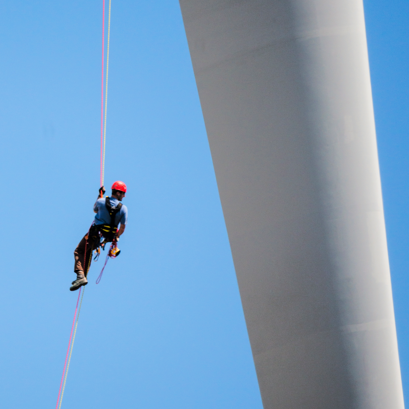 Worker hanging from cable between wind turbine rotor and tower with blue sky behind him.