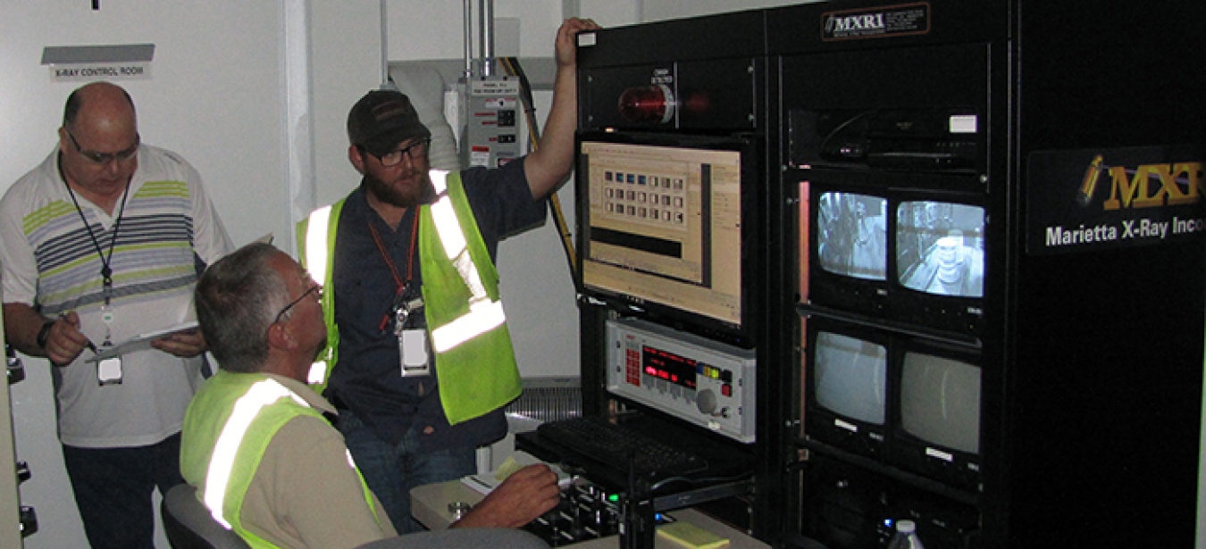 A view of the real-time radiography vault before the doors close and a waste container undergoes inspection by specialists in the adjacent control room.