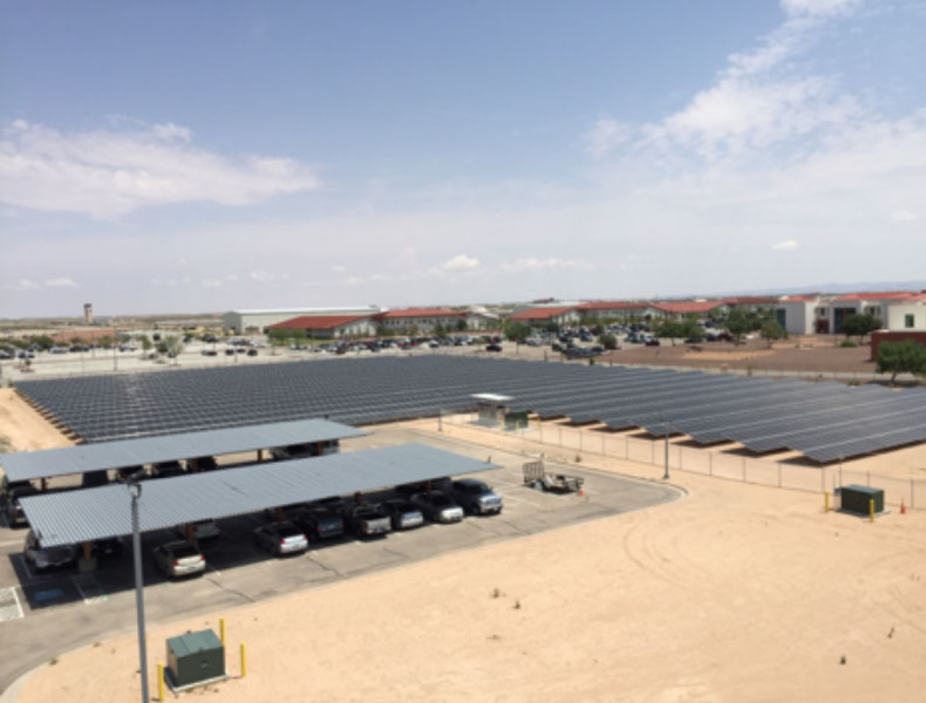 Image shows an upside down L-shaped field of dark gray solar panels on the right with a gravel lawn and a covered parking area on the left.