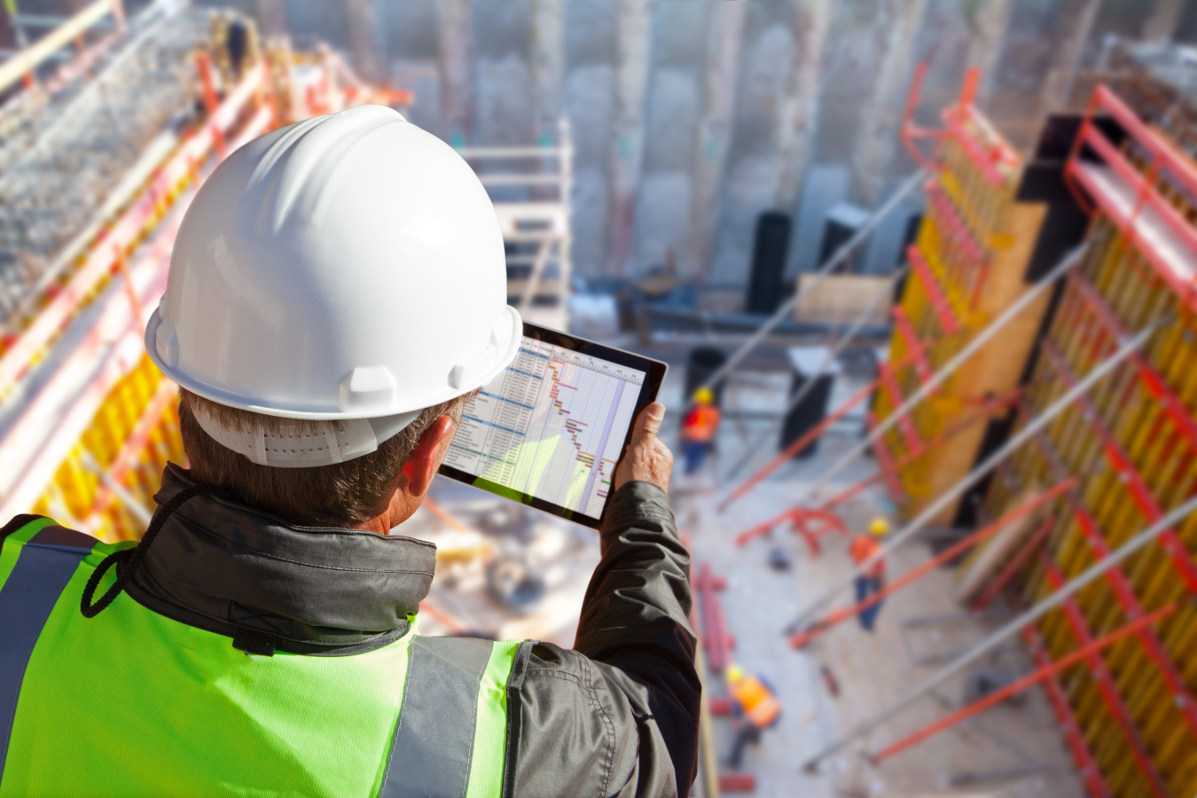 A construction worker looking at his tablet at a construction site, shown from behind him.