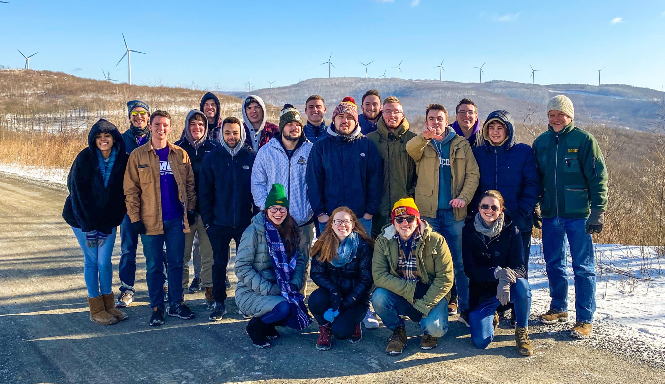 A group of young people stand in front of a wind plant.