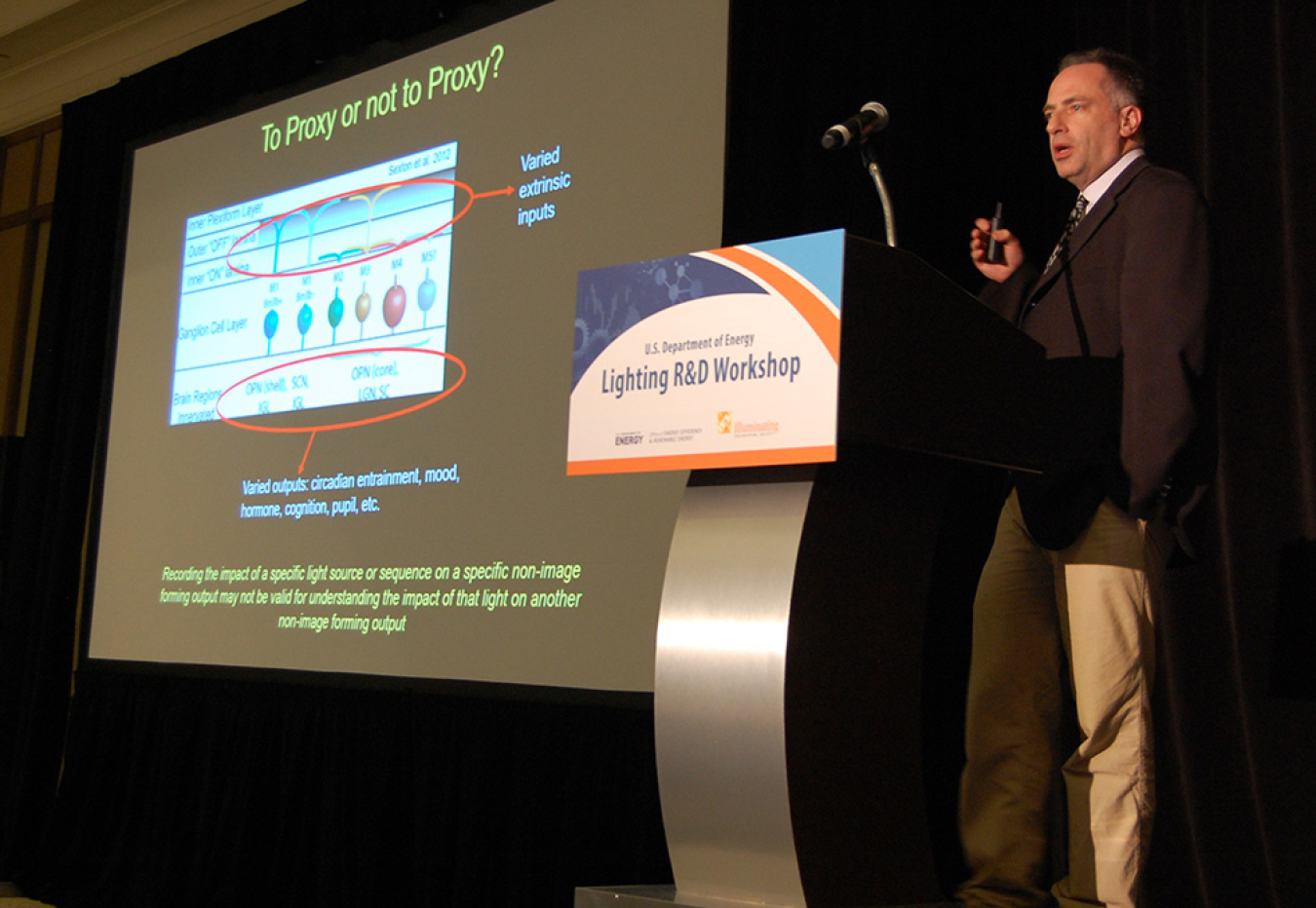 Man standing in front of the lectern addressing the conference audience, with a large projection screen next to him.