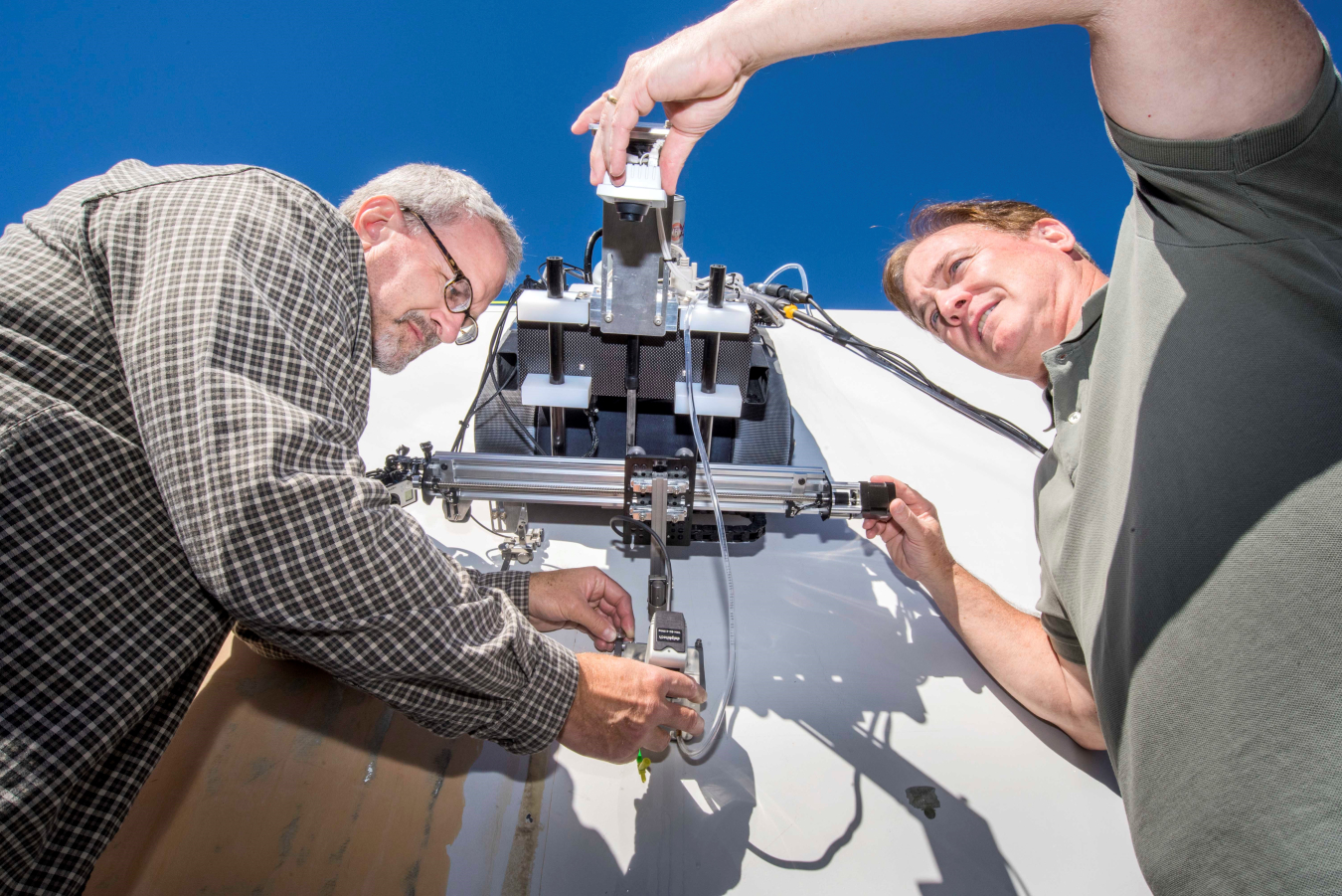 Two men set up a crawling robot against a clear blue sky.