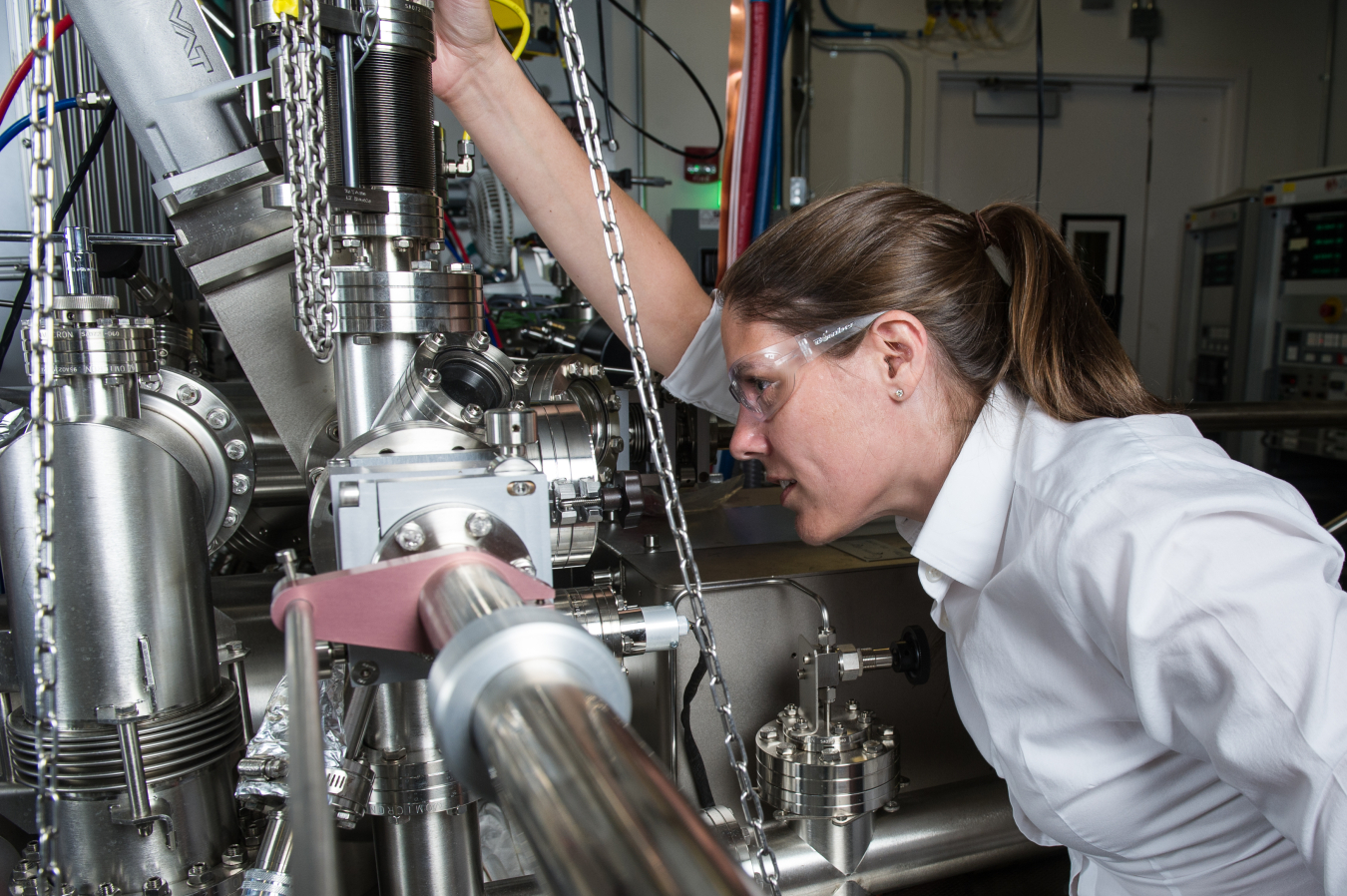 Young woman wearing protective glasses and working in a lab around heavy steel equipment.