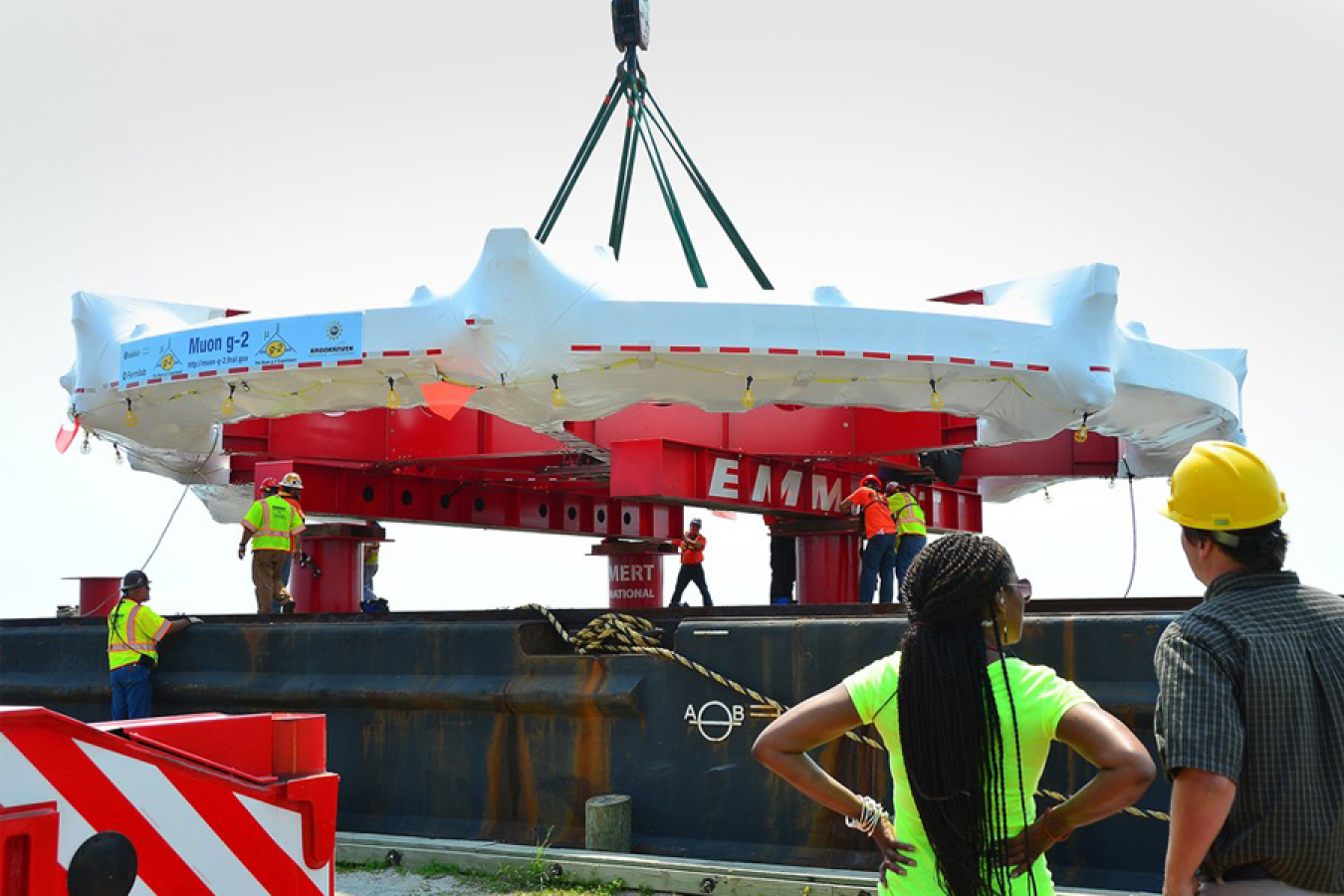 Woman and man standing with their backs to the camera looking at a giant, ring-shaped machine covered in plastic being placed on a large metal platform.