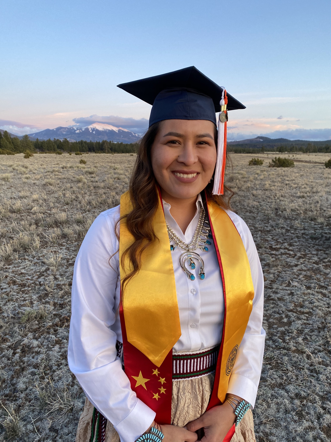 A young woman wearing a black graduation cap, red and gold graduation stole, white button-up blouse, long skirt, silver and turquoise necklace and bracelets, and woven belt stands on a field of dry grass with pine trees, mountains, and clouds behind her.