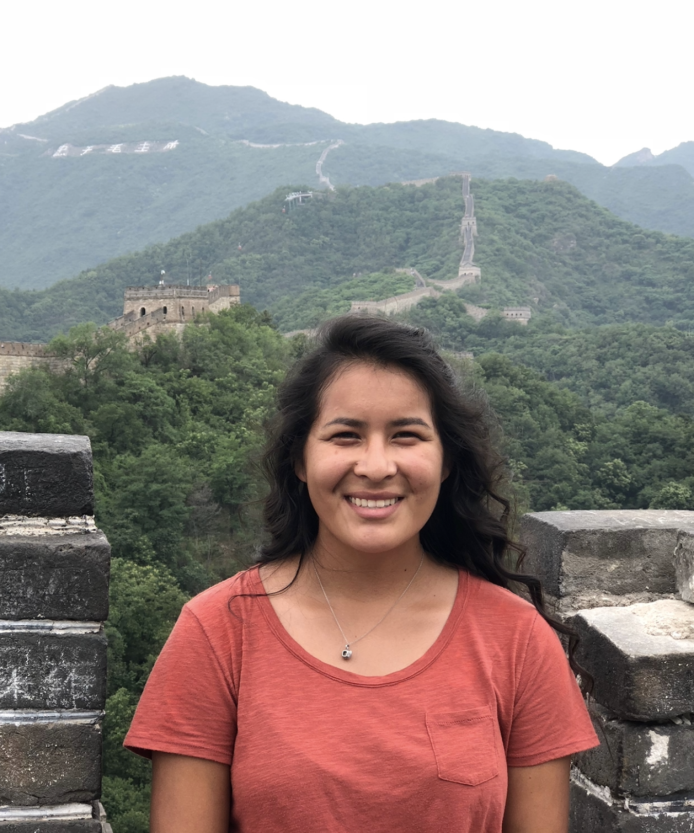 A young woman leans against a stone wall. The Great Wall of China can be seen behind her.