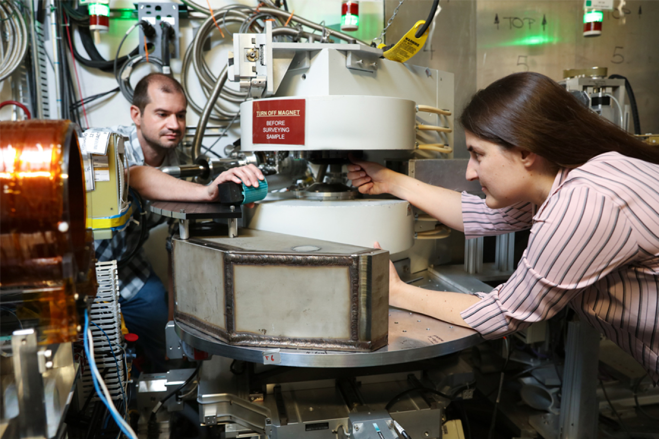 A woman and man are looking at and making adjustments to a piece of complex metal and plastic machinery with wires and tubes behind it.  