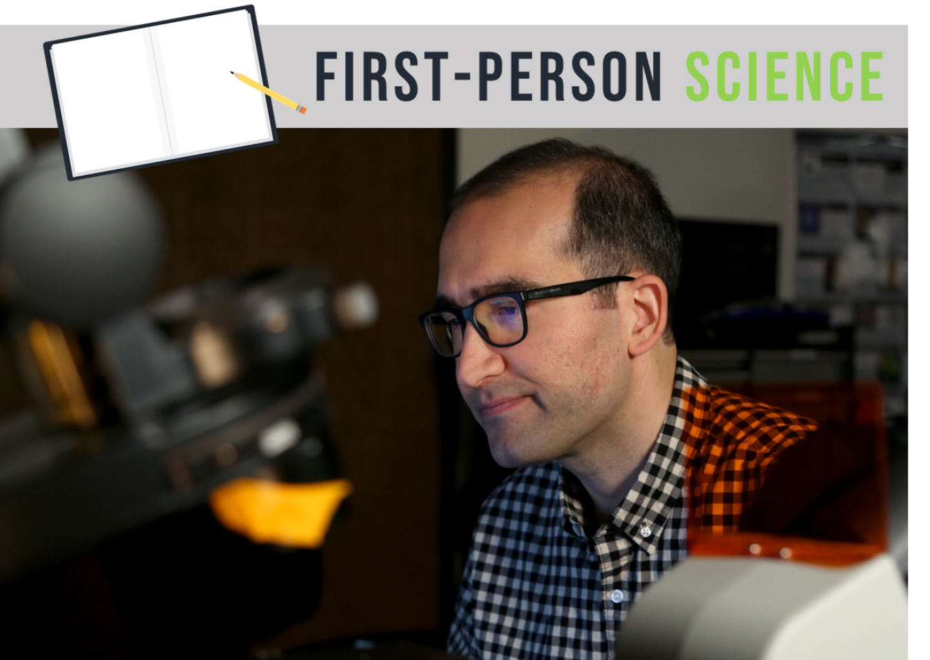 Man with a checked shirt on sitting in a laboratory next to a computer and in front of a microscope.  