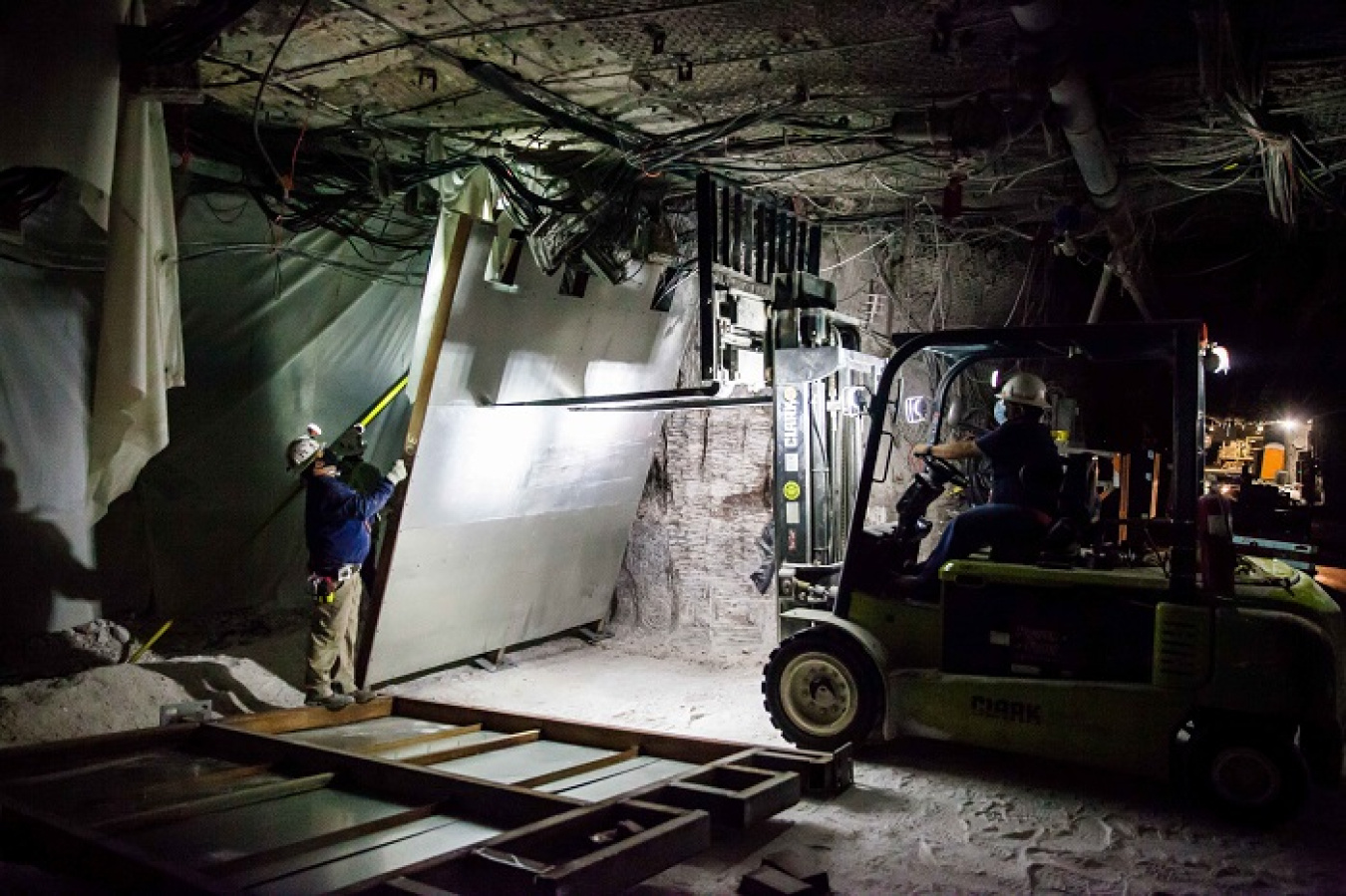 Workers in the Waste Isolation Pilot Plant underground stand up a metal bulkhead used to control airflow through one of four air circuits in the facility’s underground, 2,150 feet beneath the surface. Bulkheads are assembled in an underground fabrication shop from material transported from the surface. Foam is used to close gaps between the bulkhead and the walls.