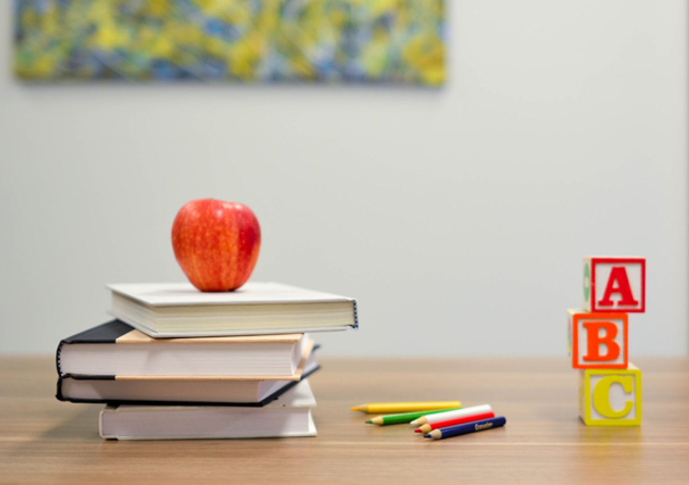 Photo of stacks of books on a table, with an apple on top, next to a few assorted-color pencils, and three building blocks with "A," "B" and "C" on them.