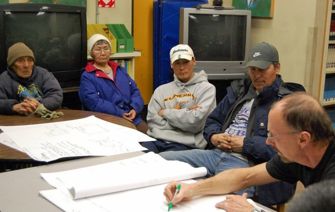 A man at right writes on a paper in the center of the table while 4 native villagers look on.th