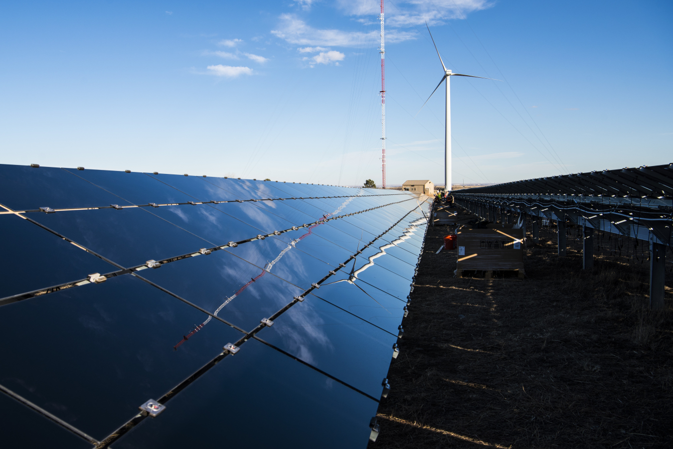 Photo of wind turbine next to a solar panel array, with images of the wind turbine and blue sky reflecting off the solar panels.