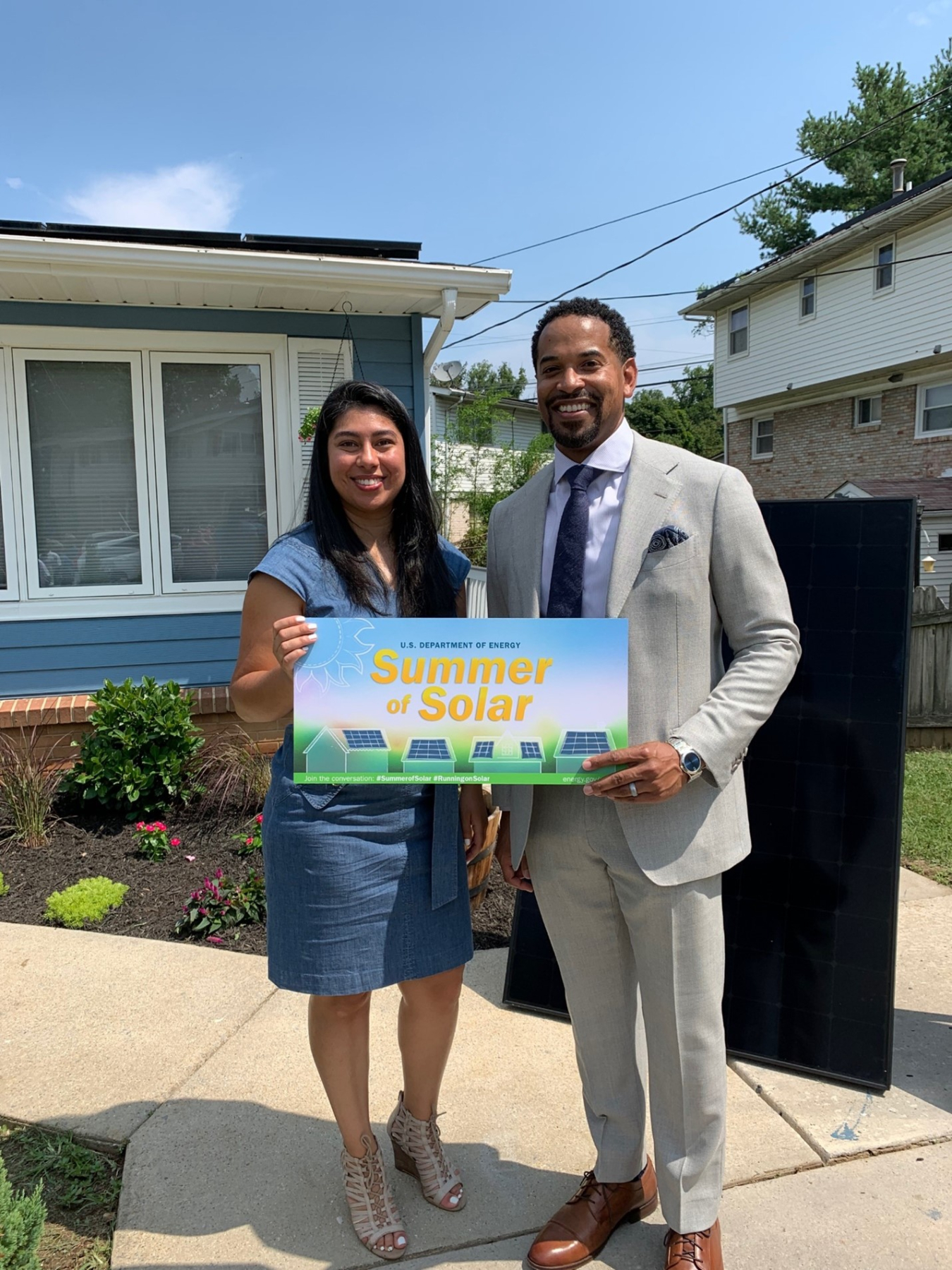 Two people standing in front of a home with solar panels holding a sign that says "Summer of Solar"