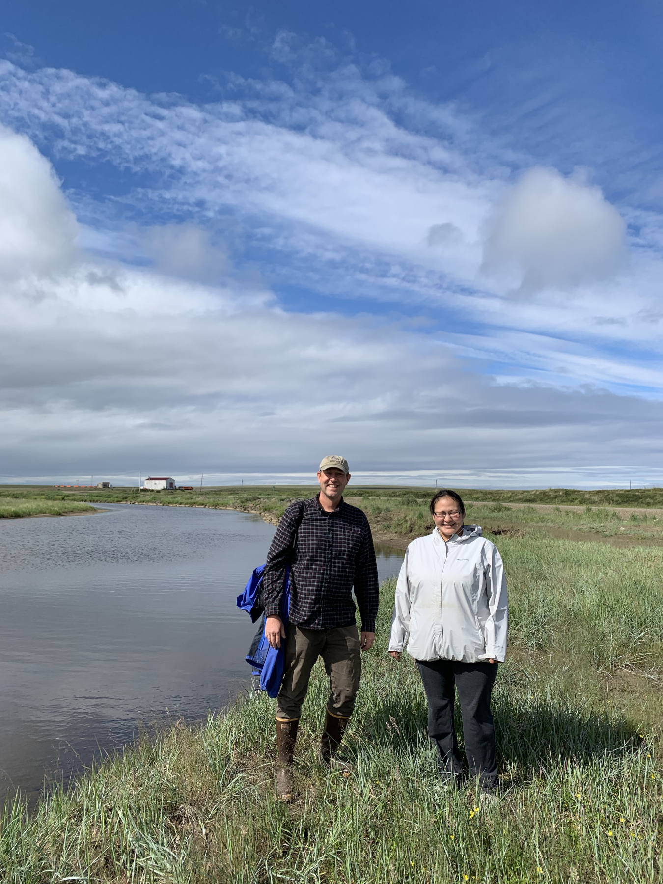 Near a river in Deering, Alaska, on a sunny day with a powerhouse in the background.