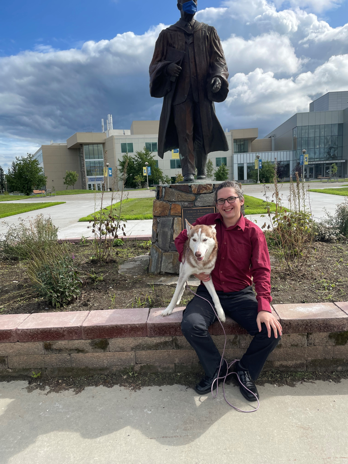 Intern Josh Hostler holds his dog and sits on a bench under a statue on the University of Alaska at Fairbanks.