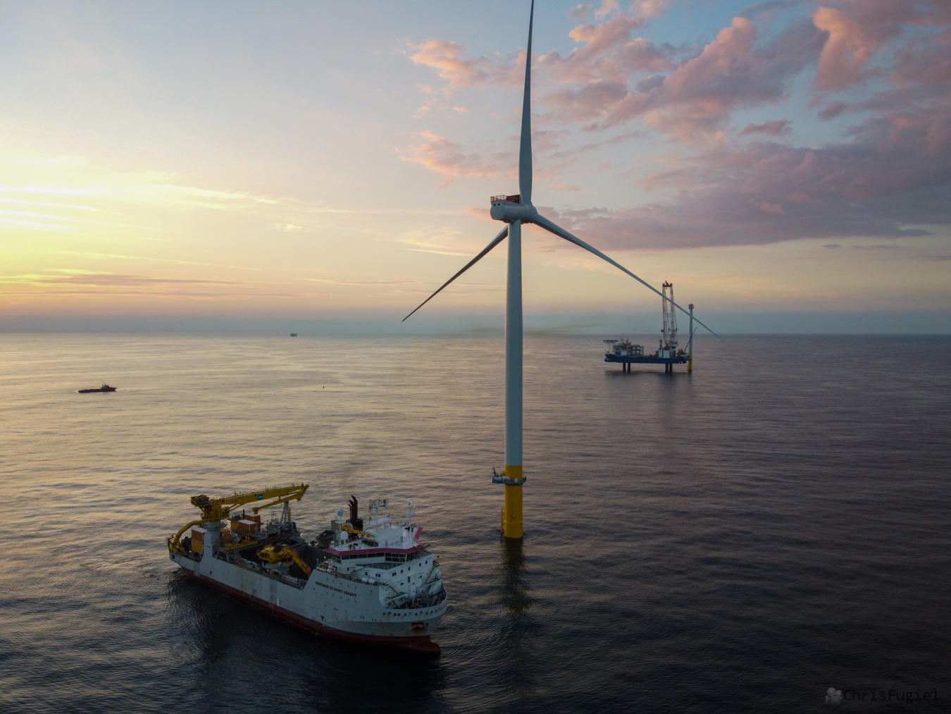 An offshore wind turbine in a calm ocean with a service vessel nearby; a platform is in the distance, erecting another wind turbine. The sun is rising (or setting) in the distance behind light clouds.