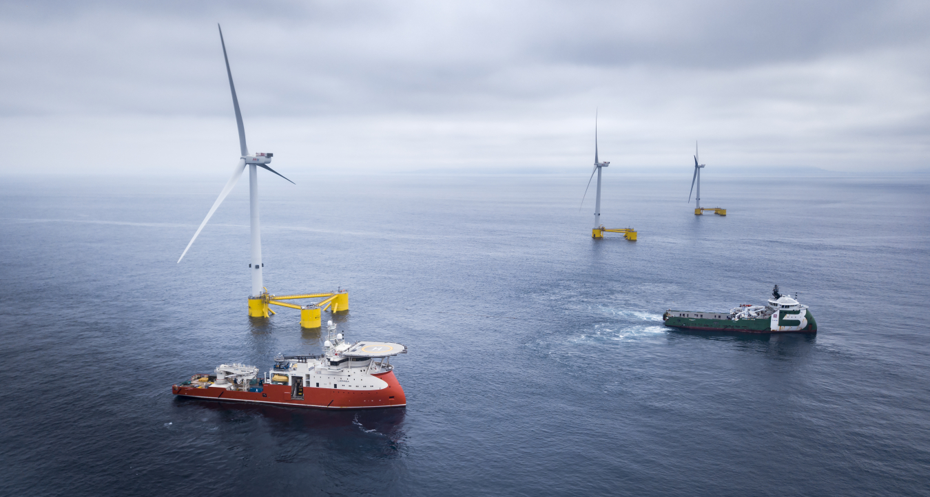 Photo of three offshore wind turbines with two ships in the foreground.