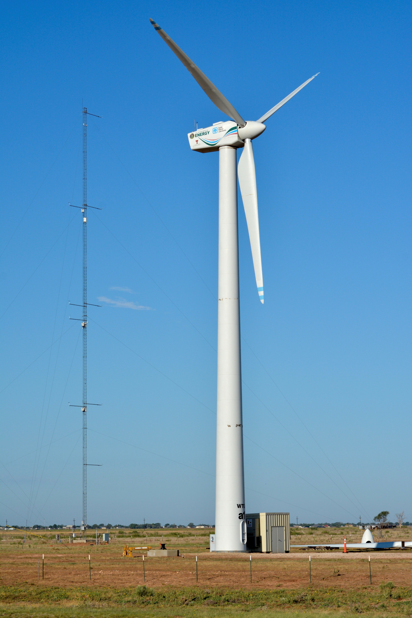 A three-bladed wind turbine installed on the ground with a meteorological tower and blue sky in the background.