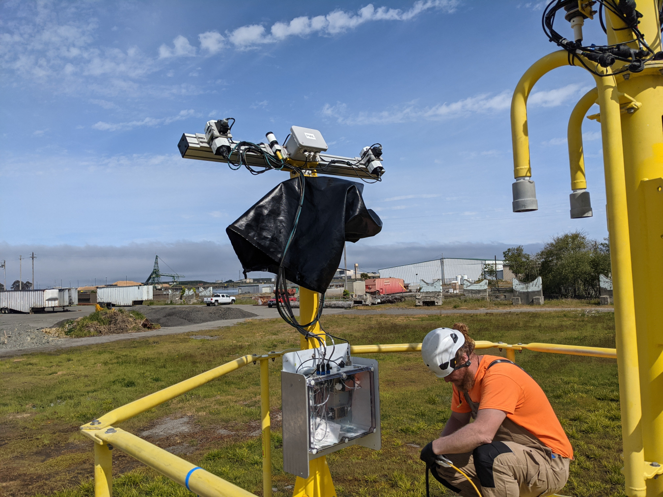 A man works on technical equipment on land. Industrial buildings and semitruck trailers are visible in the background