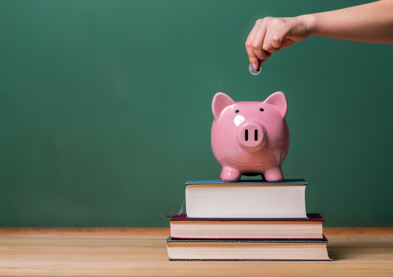 Piggy bank sitting on top of a small stack of books with a hand above it about to drop a coin in.