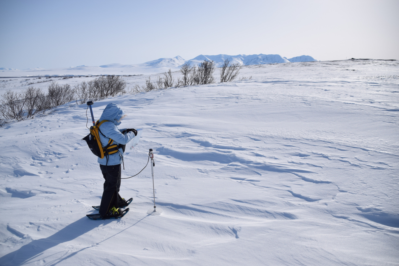 A person dressed in cold weather gear on a snow field with a scientific instrument.