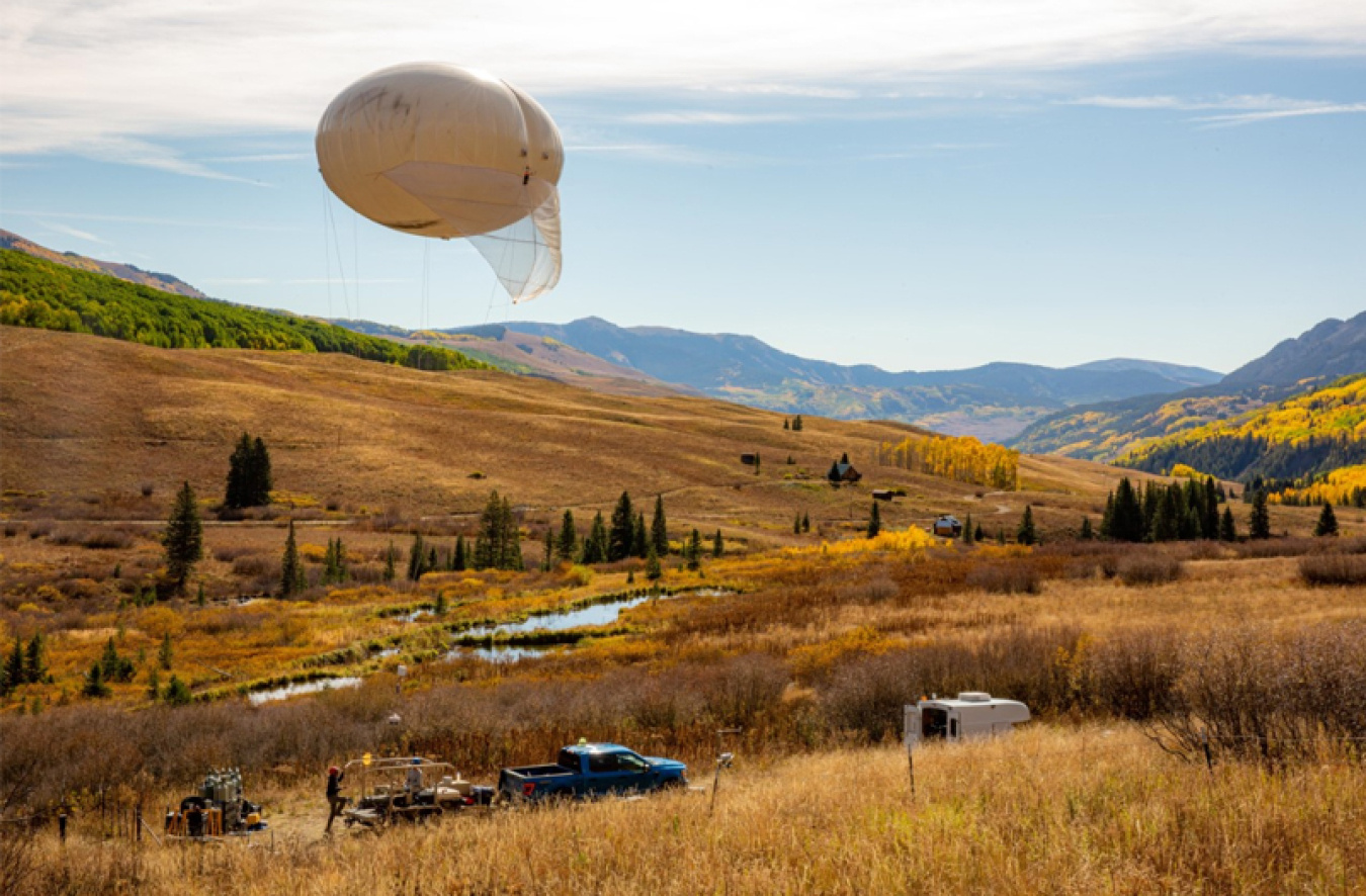 A tethered balloon attached to a truck, floats above gold fields and blue mountains