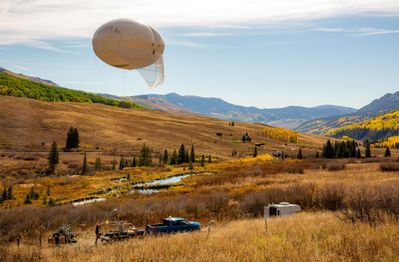 A tethered balloon attached to a truck, floats above gold fields and blue mountains 