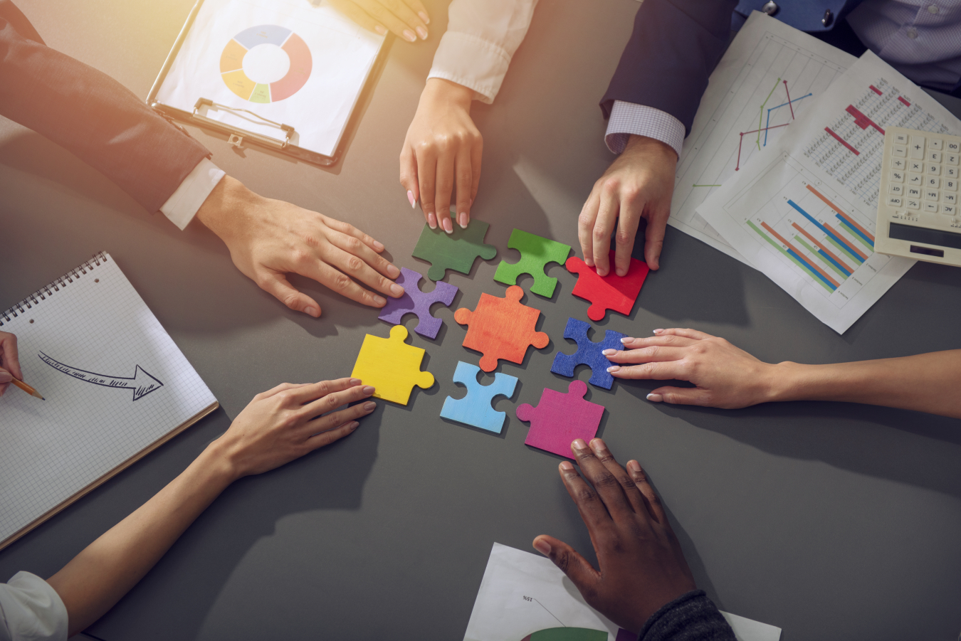 Large jigsaw puzzle pieces of different colors on a meeting table, with meeting attendee hands touching them.