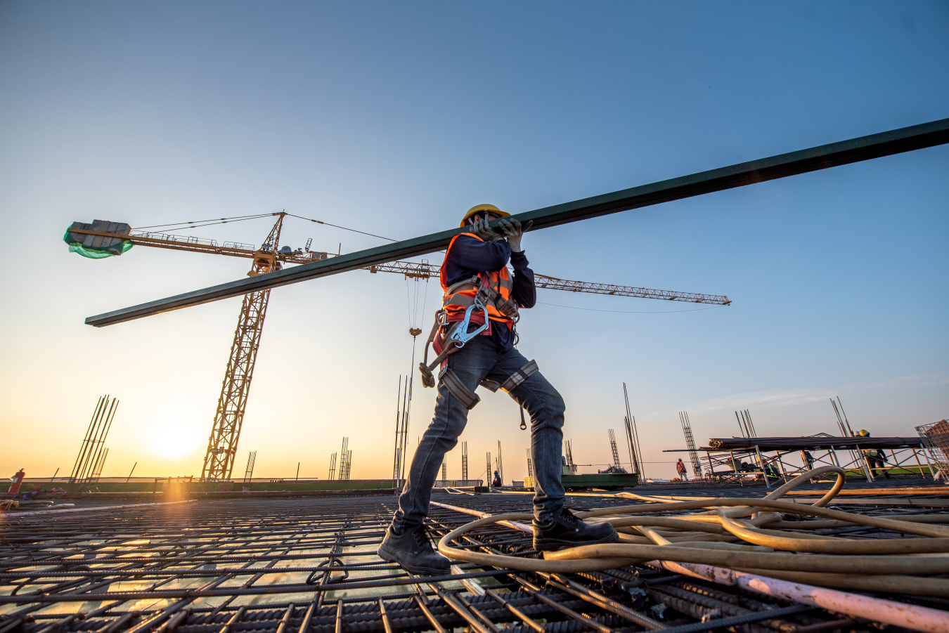 Construction worker carrying a beam across a construction site with the sun and the sky behind him.