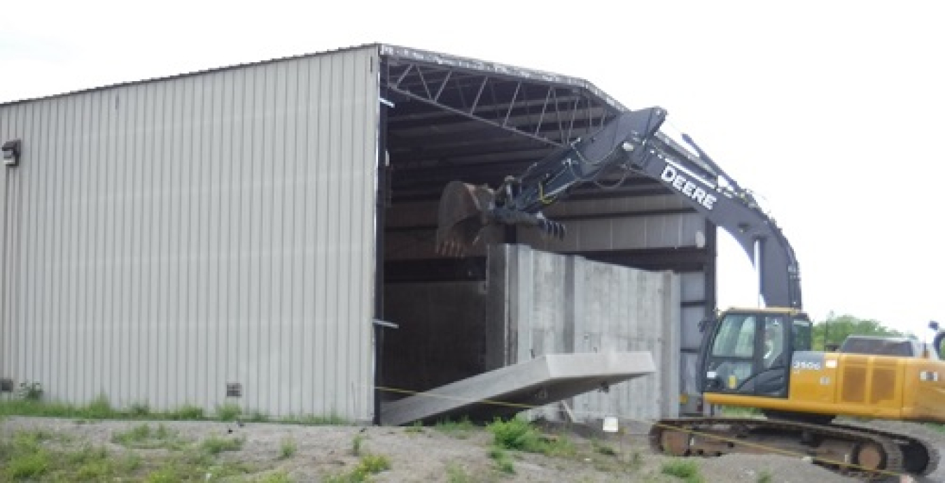 An operator at the West Valley Demonstration Project uses an excavator to remove a large concrete shield wall to create an entrance to accommodate a large fork truck for transporting empty waste containers in and out of the building.