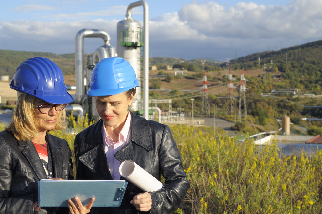 Two women wearing hard hats in front of a geothermal facility reviewing papers on a clipboard