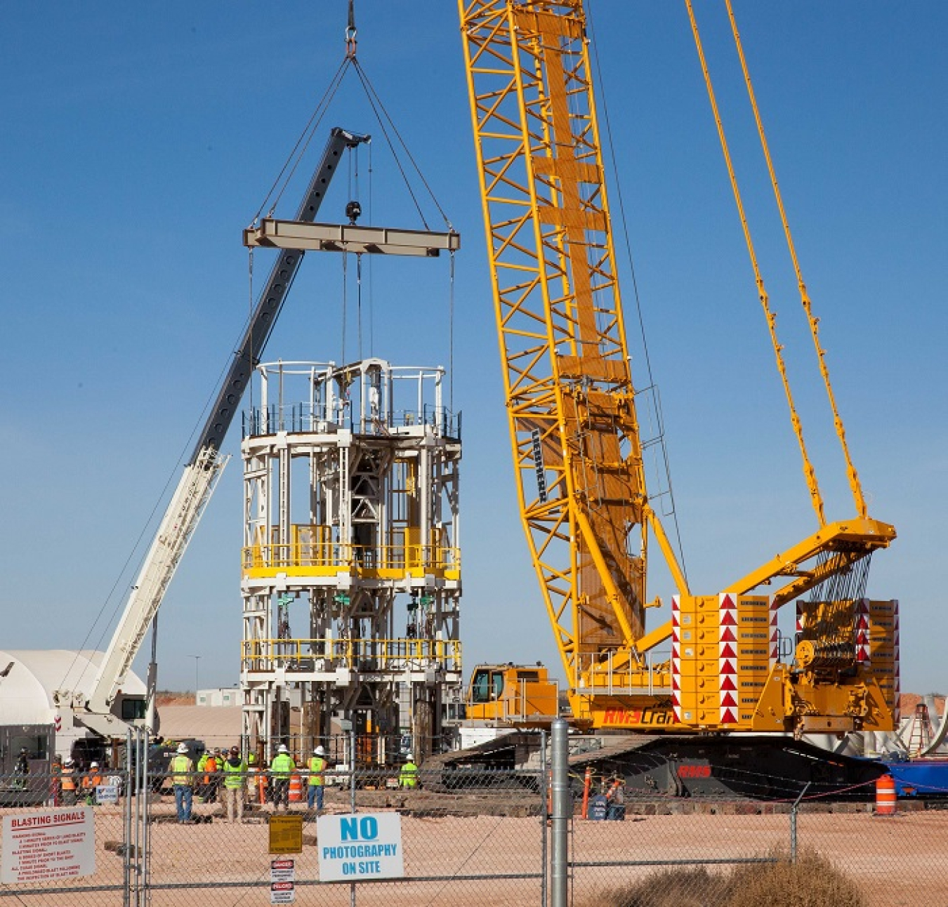 The four-story Galloway work platform is lowered into the Waste Isolation Pilot Plant’s (WIPP) utility shaft by a 550-ton crane. Excavation arms will dump rock into buckets that will be brought to the surface through a pair of holes in the platform, which allows workers to perform other tasks concurrently. When complete, the utility shaft, with a finished diameter of 26 feet, will be the fifth and largest utility shaft at WIPP.