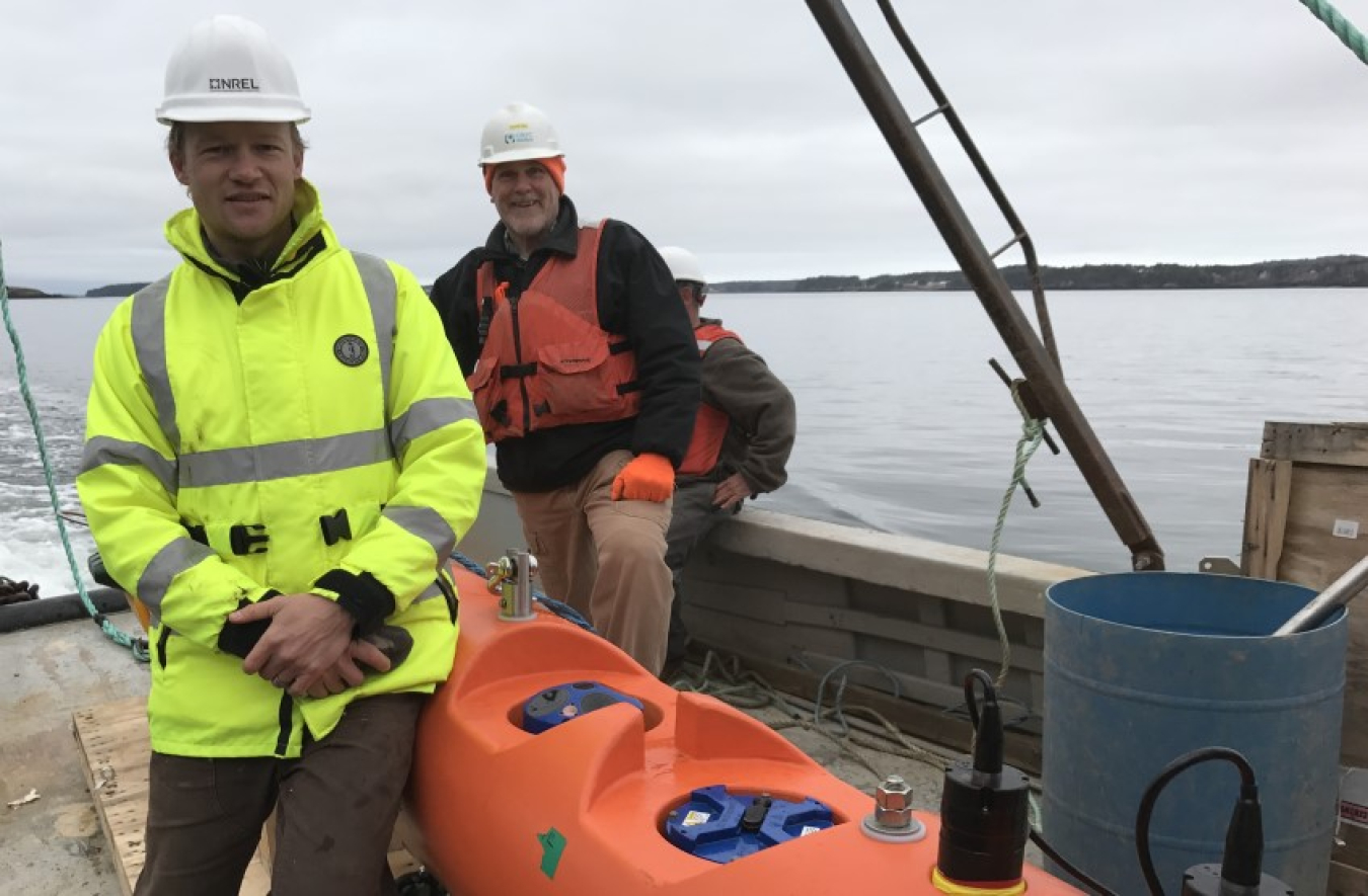 Two men in hard hats and stand next to a kayak on the deck of a boat on the water. One man is wearing a life jacket, and one is wearing a neon yellow jacket.