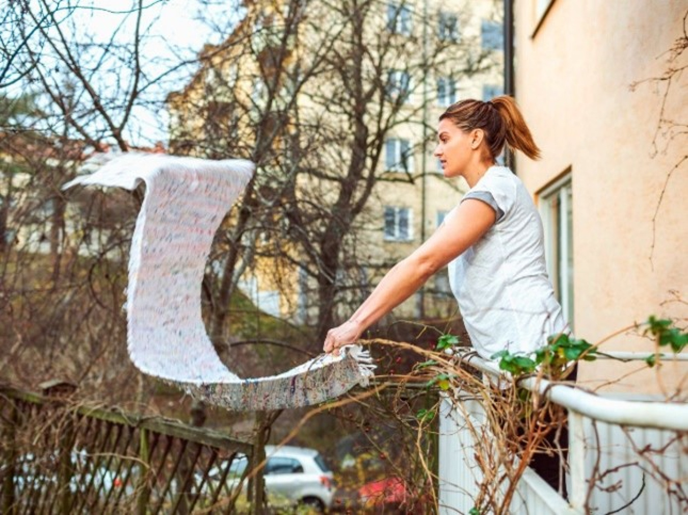 Woman standing near a railing outside a house or other building, shaking out a rug.