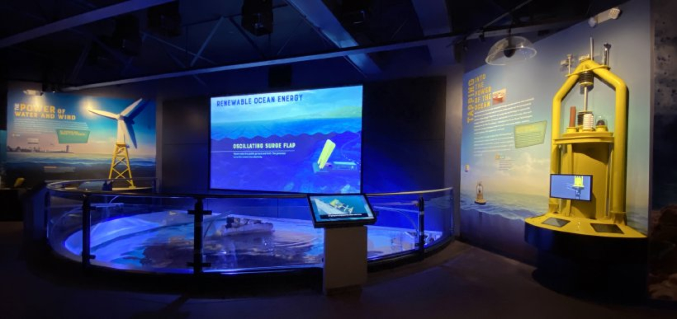 A museum exhibit with models of marine energy devices. A large screen at the center of the exhibit displays animations of different marine energy technologies.