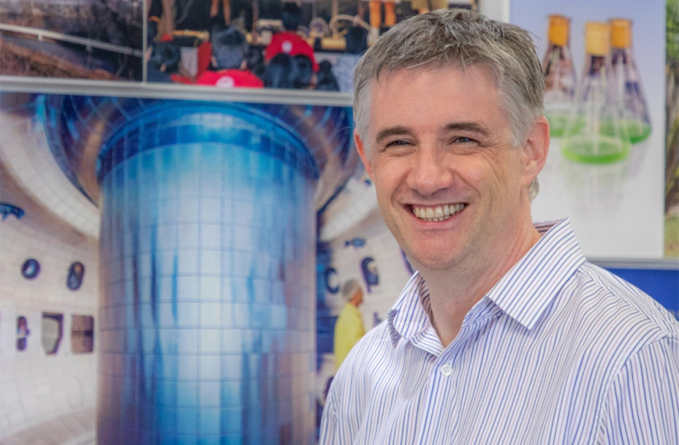 Richard Buttery, Director of the DIII-D National Fusion Facility, stands in front of an image showing the interior of the DIII-D tokamak.