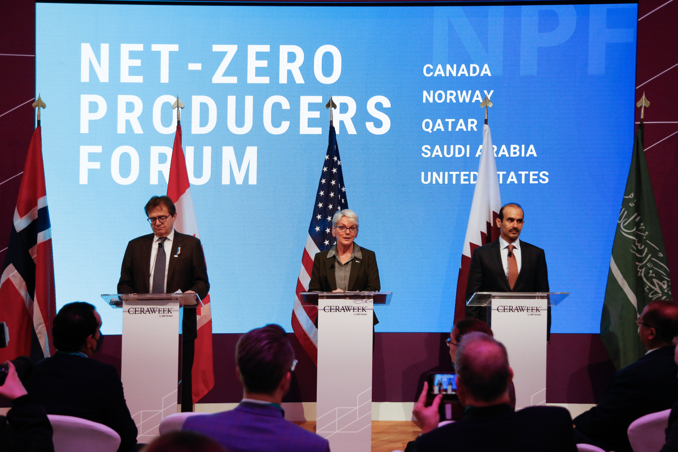 Secretary of Energy Jennifer Granholm stands in the middle of three people at podiums in front of a crowd. In the background are the words "Net-Zero Producers Forum" in white lettering against blue on a large screen.