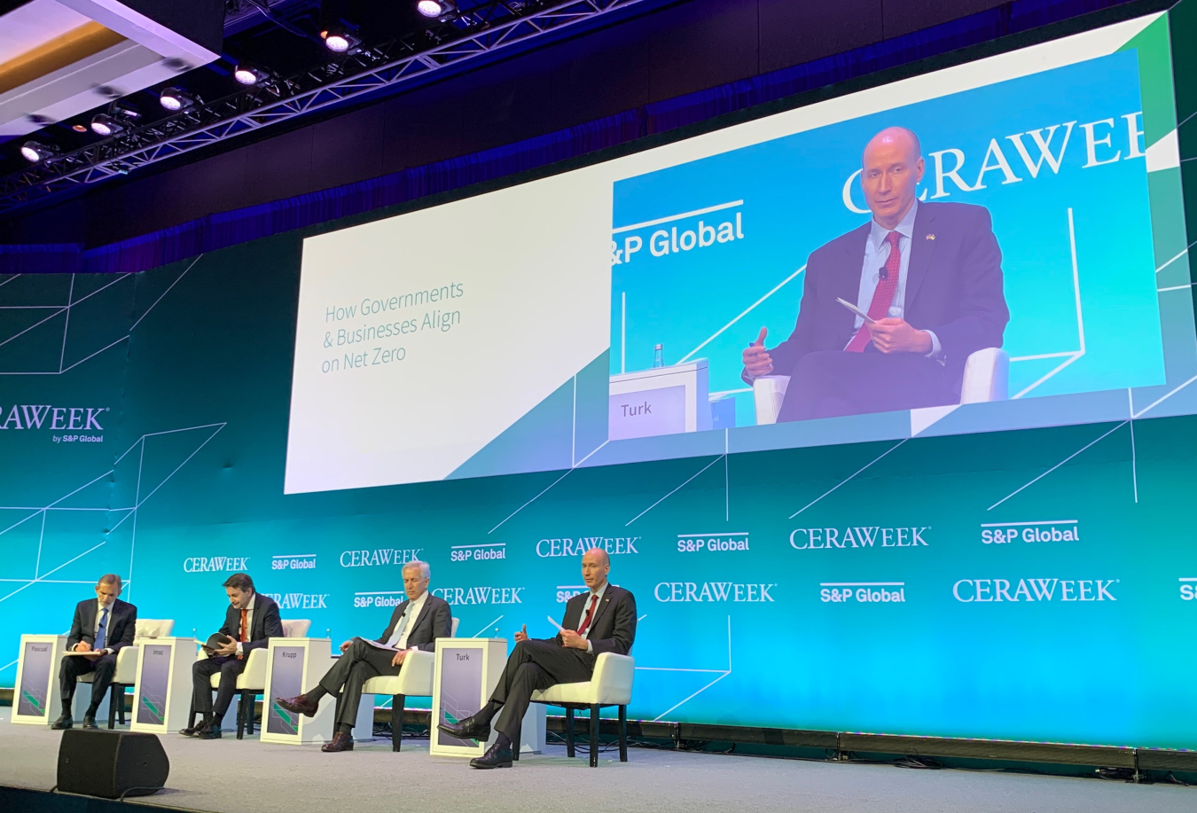 An image taken from far away of the Deputy Secretary sitting in a white chair on a grey stage with three other people. In the background there is a teal panel containing the CERAWeek and S&P Global logos and a large screen that shows the participants.