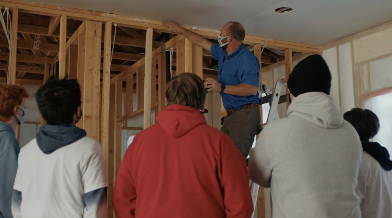 Small group of people in a room, their backs to the camera, with one man up on a ladder examining the interior wall construction of the room.