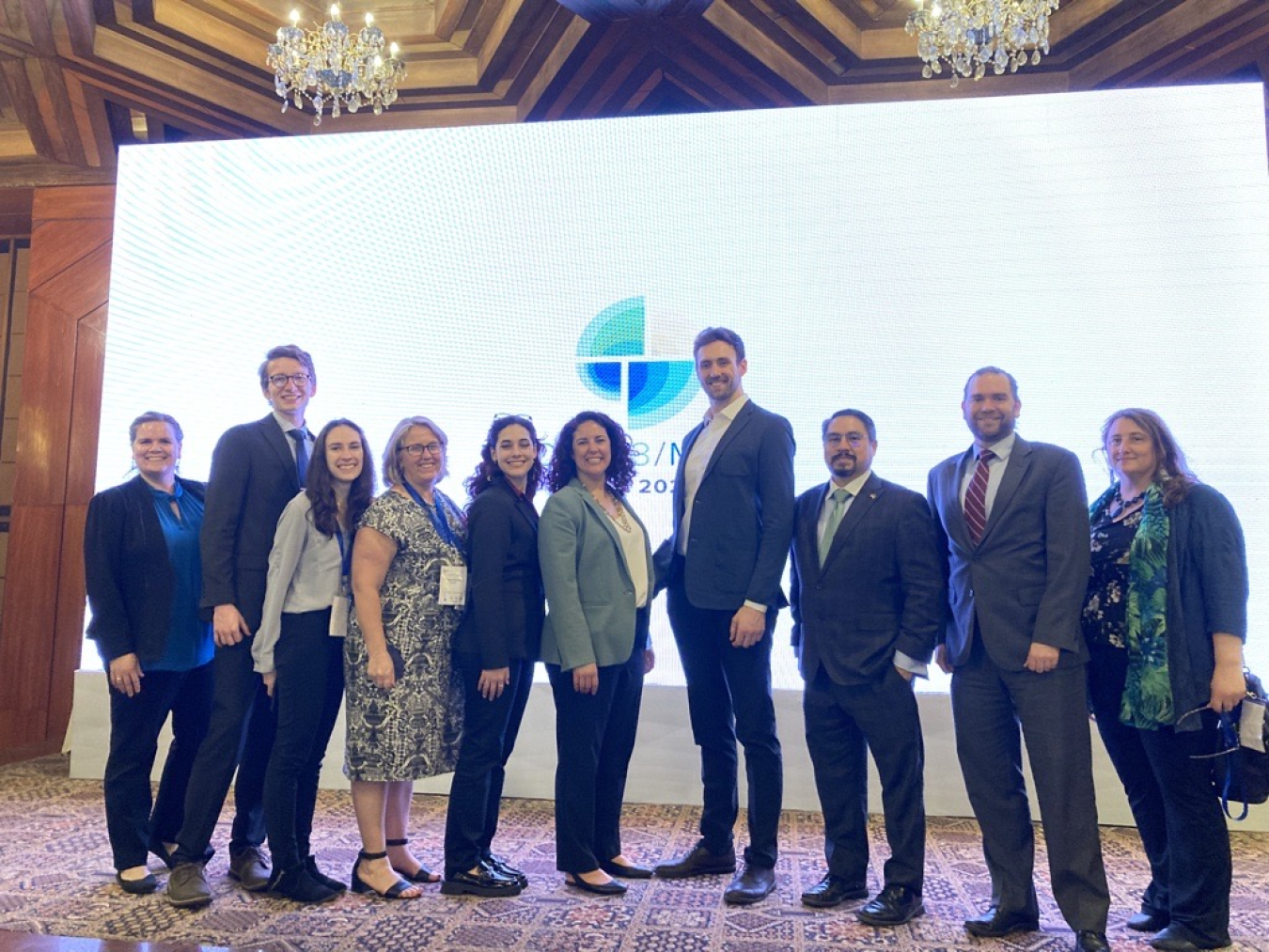 A group of representatives from the Office of International Affairs stand in front of a white screen showing the yellow, blue and green Clean Energy Ministerial and Mission Innovation logo in a hotel in New Delhi, India.