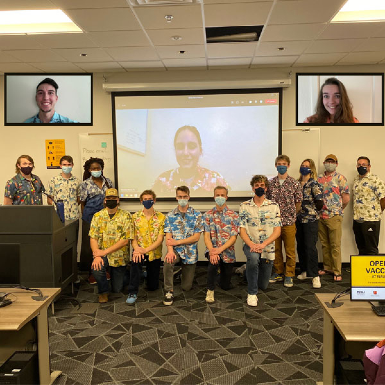 Members of the WindJax Energy Club standing at the front of a classroom wearing medical masks and Hawaiian shirts. A few students are projected on screens behind them. 