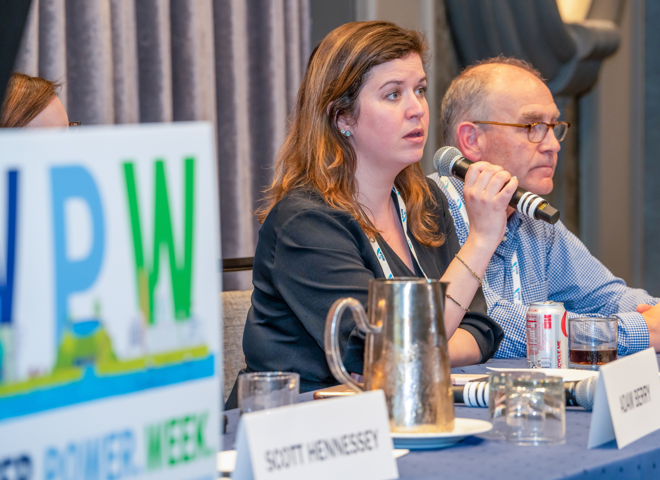 Two people are sitting side by side at a table. One person is speaking into a microphone. 